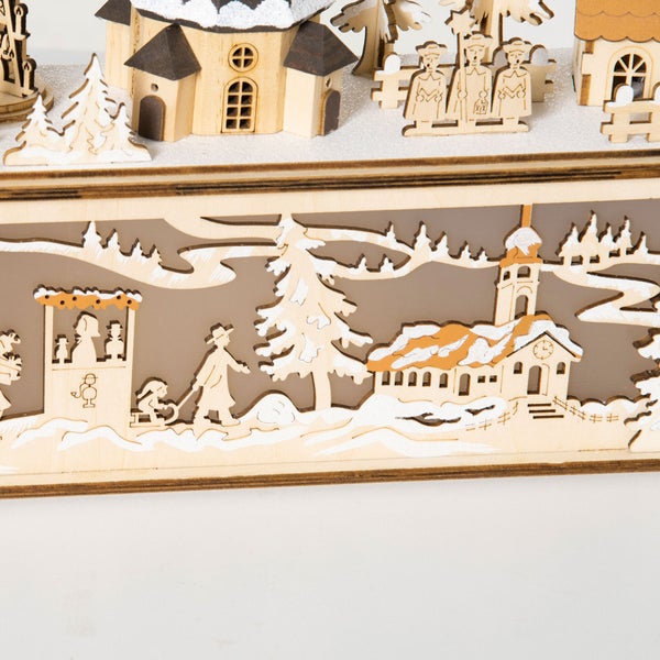 Nahaufnahme einer hölzernen Weihnachtsdekoration mit einem lasergeschnittenen Winterdorf-Motiv, Häusern, Figuren und verschneiten Bäumen.