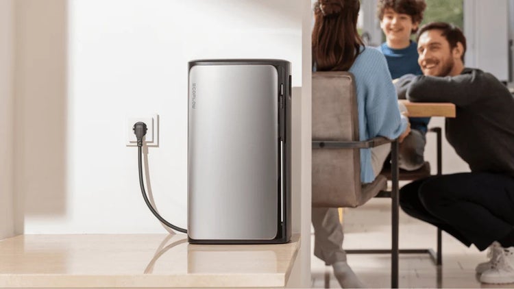 EcoFlow tragbare Powerstation in Silber und Schwarz, an einer Wandsteckdose angeschlossen, in einem hellen Raum mit einer Familie im Hintergrund.