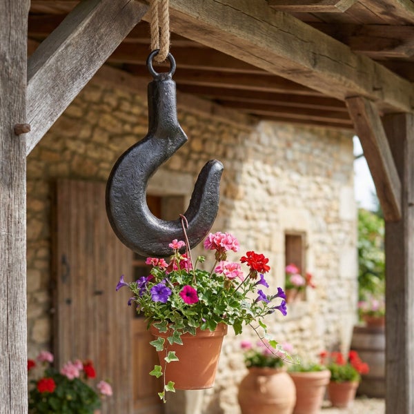 Ein rustikaler Blumenampelhalter in Form eines Kranhakens aus Metall, der unter einem hölzernen Vordach hängt und einen Tontopf mit Blumen hält.