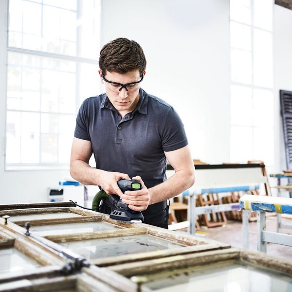 Mann schleift Holzfenster mit einem Exzenterschleifer. Der Schleifer ist an eine Staubabsaugung angeschlossen.