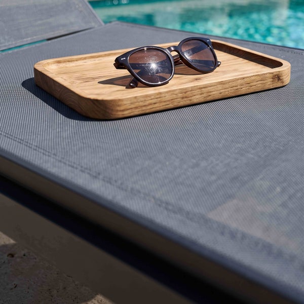 Holztablett mit Sonnenbrille auf einer Gartenliege mit Netzgewebe an einem Swimmingpool.
