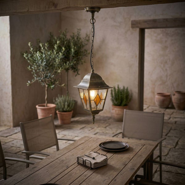 Rustikale Außenhängeleuchte aus Metall und Glas über einem hölzernen Gartentisch auf einer mediterranen Terrasse mit Pflanzen.