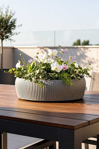 Runde, geriffelte Pflanzschale auf einem Holztisch, bepflanzt mit weißen und rosa Blumen sowie Efeu auf einer hellen Terrasse.