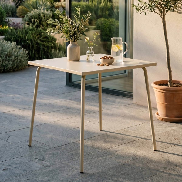 Quadratischer Bistrotisch aus Metall auf einer Steinterrasse, dekoriert mit Vasen, einer Schale Nüsse und einem Wasserkrug in hellem Gartenambiente.