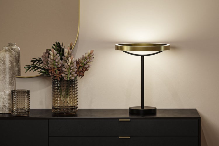 Moderne Tischleuchte mit ringförmigem goldenem Schirm auf einer dunklen Kommode neben einer Blumenvase und einem Spiegel.