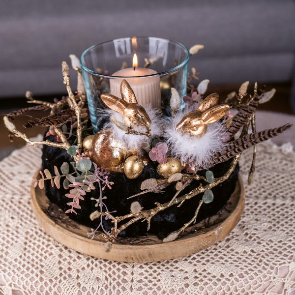 Ostergesteck mit zwei Hasenfiguren, Kerze im Glas, Zweigen und Federn auf einem runden Holztablett, frühlingshafte Tischdekoration.