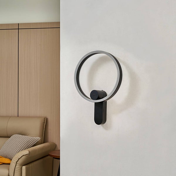 Moderne LED-Wandleuchte in Ringform mit schwarzer Halterung an einer hellen Wand neben einem beigen Sofa.