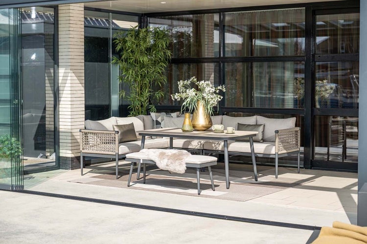 Moderne Garten-Lounge-Gruppe mit Ecksofa, Esstisch und Sitzbank. Hellgraue Polster und Seilgeflecht-Details auf einer hellen, überdachten Terrasse.