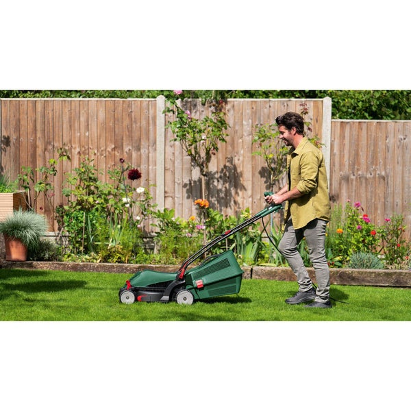 Ein Mann mäht den Rasen mit einem grünen elektrischen Rasenmäher in einem hellen, organisierten Garten mit Holzzaun und Blumen.