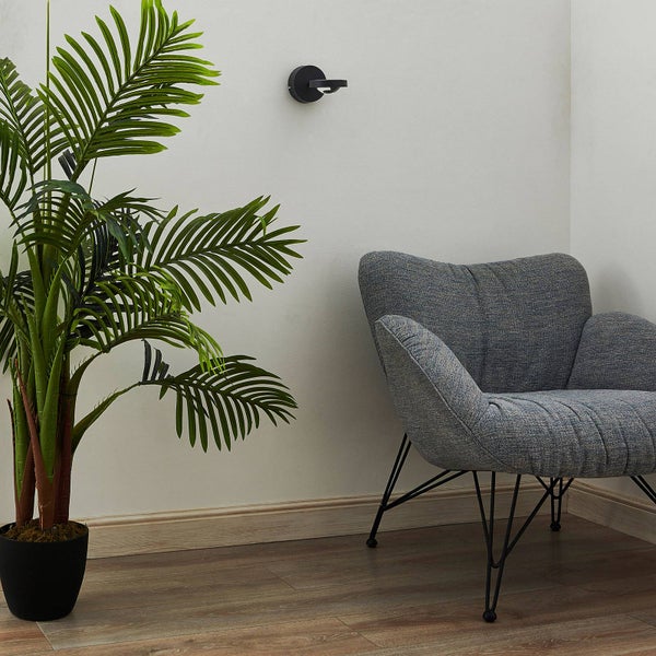 Kleine schwarze Wandleuchte über einem grauen Polstersessel in einem modernen Raum mit einer großen künstlichen Palme.