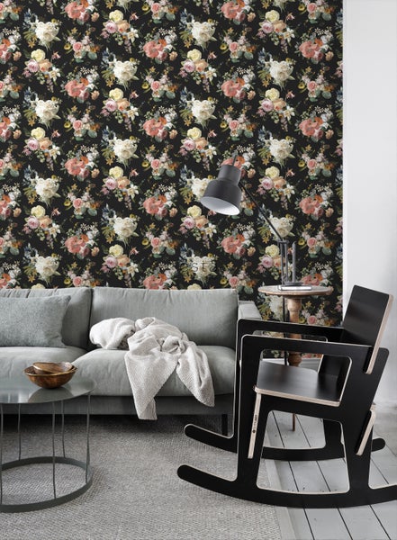 Ein Wohnzimmer mit einer Wandtapete mit Blumenmuster, einem grauen Sofa und einem schwarzen Schaukelstuhl.