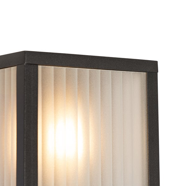 Detailansicht einer Außenwandleuchte mit dunklem Metallrahmen und vertikal geripptem Milchglas mit eingeschaltetem Leuchtmittel.