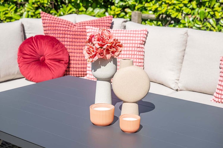 Moderner Gartentisch mit dekorativen Vasen, Kunstblumen, Windlichtern und Loungesofa mit Kissen im hellen Außenbereich.