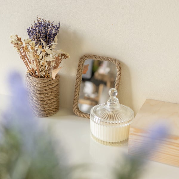 Kleiner Wandspiegel mit Juteseil-Rahmen, Glas-Duftkerze und getrocknete Blumen in Jute-Vase auf heller Ablage.