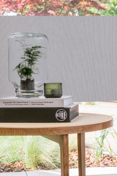 Lichtdurchlässiges graues Rollo mit vertikalem Streifenmuster hinter einem runden Holztisch mit Pflanzenglas und Büchern.