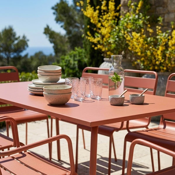 Moderner Gartentisch mit passenden Stühlen in Terrakotta, gedeckt mit Keramikgeschirr und Gläsern auf einer hellen Terrasse.