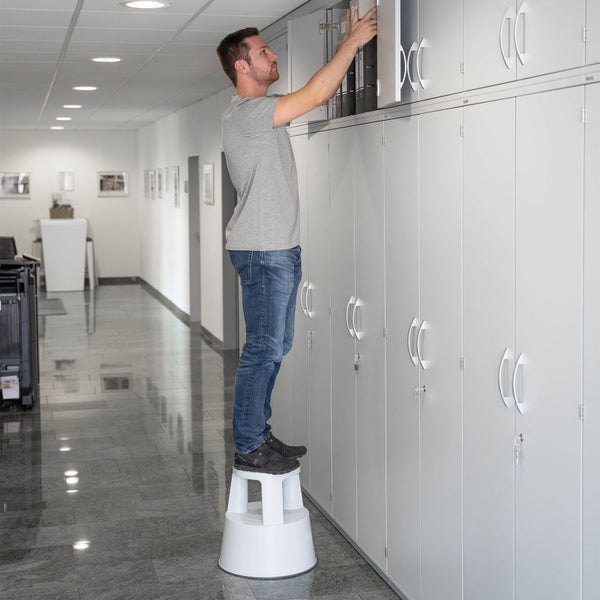 Ein Mann steht auf einem Tritthocker und räumt Ordner in einen Aktenschrank in einem Büro.