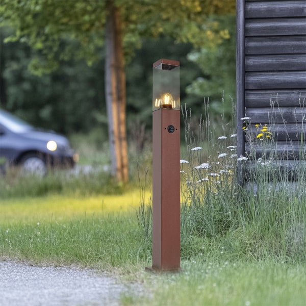 Moderne eckige Wegeleuchte aus Metall in Rostoptik mit Glasgehäuse und Sensor an einem Gartenweg neben einer Holzwand.