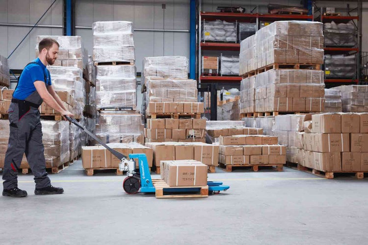 Ein Mann bedient einen blauen Handhubwagen in einem Lager mit gestapelten Paletten und Kartons.