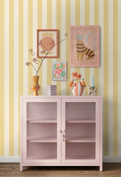 Vliestapete mit vertikalen Streifen in Gelb und Weiß hinter einem rosa Schrank mit Glasfront und Dekoration.