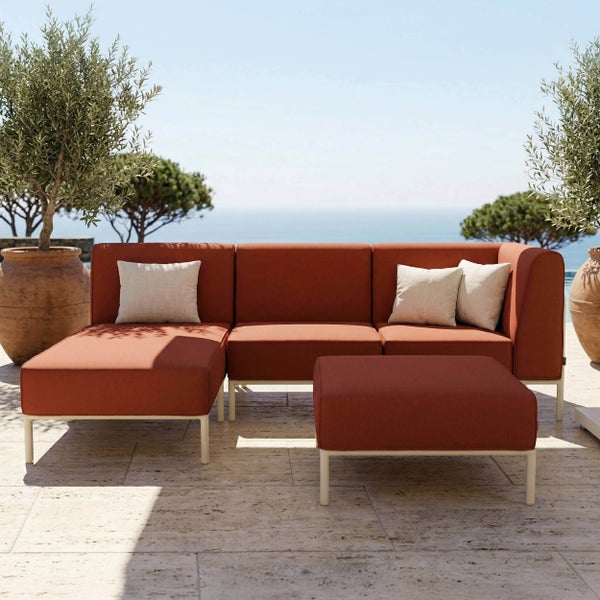 Modulare Gartenlounge mit rostfarbenen Polstern und hellem Metallgestell auf einer sonnigen Steinterrasse mit Meerblick.