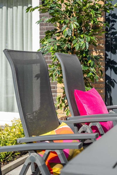 Gartenstühle mit Textilbespannung und bunten Kissen auf einer Terrasse vor einer Ziegelwand und Pflanzen.