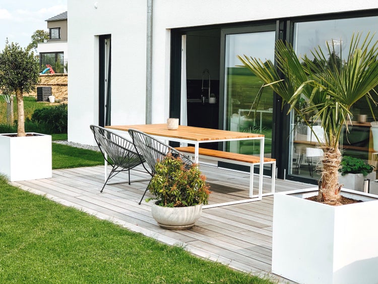 Moderne Terrasse mit Holztisch, Gartenbank und zwei schwarzen Loungesesseln auf einem Holzdeck neben einer Rasenfläche.