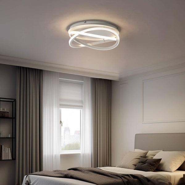 Moderne LED-Deckenleuchte im verschlungenen Ringdesign in einem hellen Schlafzimmer mit Bett und grauen Vorhängen.
