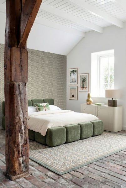 Helles Schlafzimmer mit grünem Polsterbett, gemusterter Tapete, Teppich und einem rustikalen Holzbalken auf Steinboden.