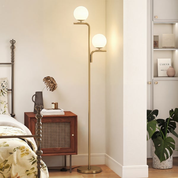 Eine moderne goldene Stehlampe mit zwei weißen kugelförmigen Glasschirmen steht in einem hellen Schlafzimmer neben einem Nachttisch aus Holz.