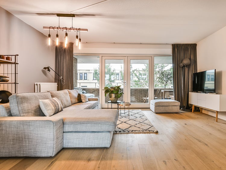 Modernes, helles Wohnzimmer mit großem grauen Ecksofa aus Stoff, hellem Echtholzboden, Glastüren zum Balkon und dekorativer Pendelleuchte.