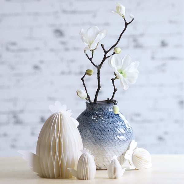 Helle Frühlingsdekoration mit blauer Keramikvase, Magnolienzweigen und Papier-Wabenfiguren von Huhn und Hase auf einem Holztisch.