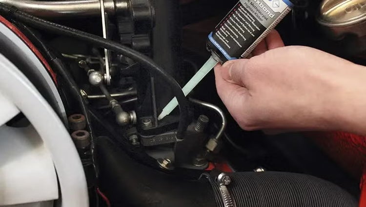 Eine Hand trägt Hightech Kleber aus einer Tube auf ein Bauteil im Motorraum eines Kraftfahrzeugs auf.