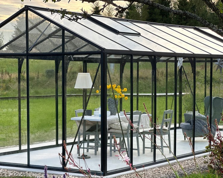 Modernes Glasgewächshaus mit Metallrahmen, eingerichtet als Gartenzimmer mit Tisch, Stühlen und Stehlampe in einem Garten.