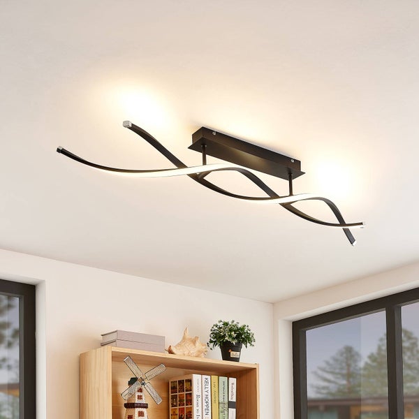 Moderne schwarze LED-Deckenleuchte mit geschwungenen Lichtarmen in Wellenform, montiert in einem hellen Zimmer über einem Holzregal.