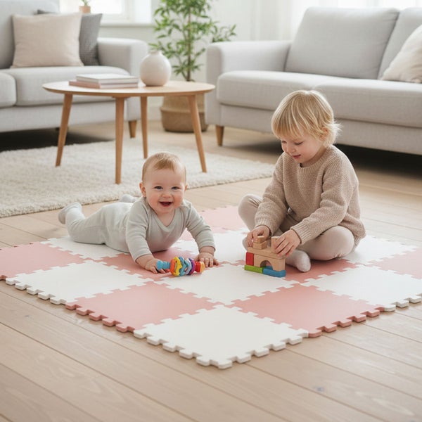 Helles Wohnzimmer mit zwei Kindern, die auf einer quadratischen weiß-rosa Puzzle-Spielmatte aus Schaumstoff auf einem Holzboden spielen.
