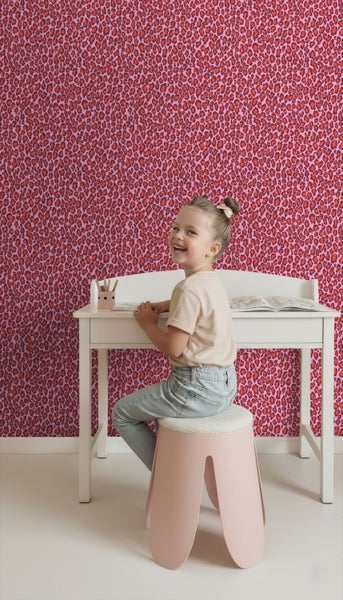 Helles, organisiertes Kinderzimmer mit weißem Schreibtisch vor einer Wand mit rot-rosa Leoparden-Tapete und einem sitzenden Kind.