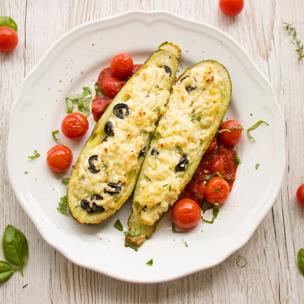 Zwei gefüllte Zucchini-Hälften mit Käse und schwarzen Oliven auf Tomatensauce mit Kirschtomaten serviert.