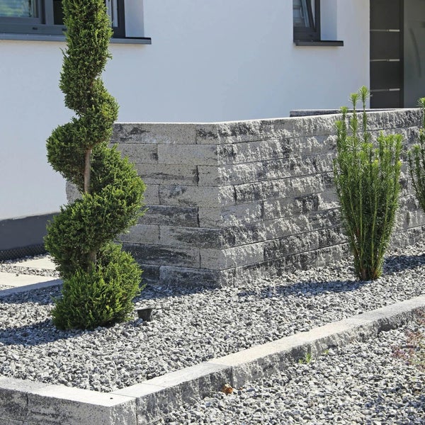 Gartengestaltung mit einer modernen Mauer aus Betonstein, ergänzt durch einen spiralförmig geschnittenen Lebensbaum und Kiesbeet vor einem Haus.