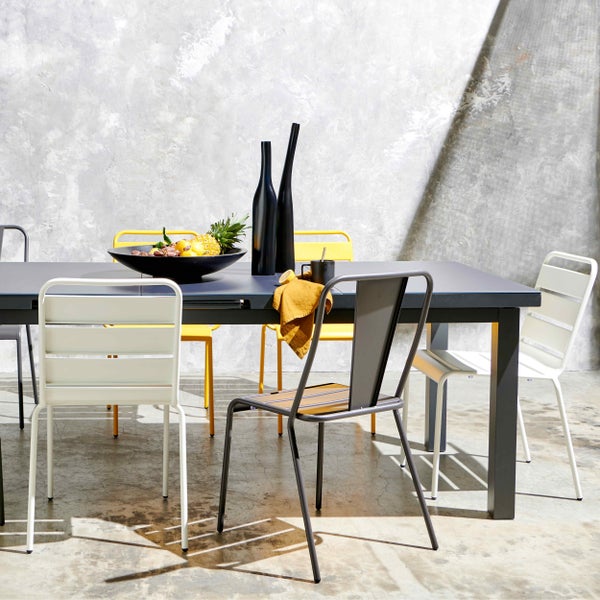 Moderner Essbereich mit dunklem rechteckigem Tisch und Metallstühlen in Weiß, Gelb und Grau vor einer Wand in Betonoptik.