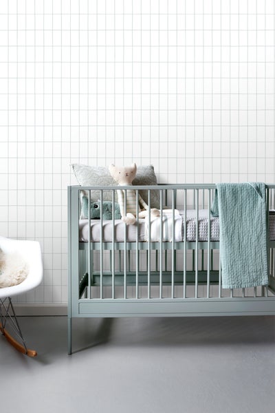 Vliestapete mit Karomuster an der Wand in einem hellen Kinderzimmer mit einem Kinderbett und einem Schaukelstuhl.