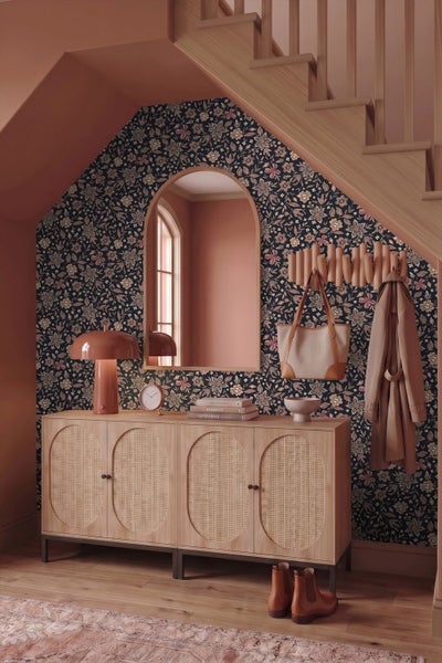 Flur mit Sideboard aus Holz und Wiener Geflecht, floraler Tapete, Bogenwandspiegel und Holztreppe.
