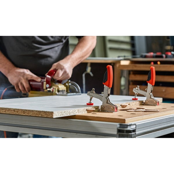 Ein Mann benutzt eine Oberfräse auf einem Holzbrett, das mit Knebelspannern auf einem Tisch befestigt ist.