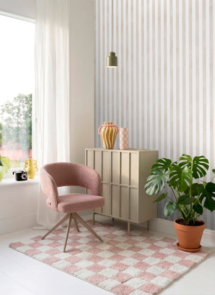 Helles Interieur mit vertikal gestreifter Tapete, rosa strukturiertem Drehsessel, rosa-weiß kariertem Teppich und beigem Sideboard aus Holz.
