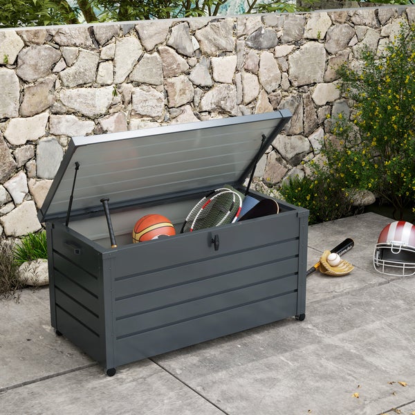 Große dunkelgraue Gartenbox aus Metall mit geöffnetem Deckel, befüllt mit Sportgeräten wie Basketbällen und Tennisschlägern vor einer Steinmauer.