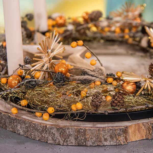 Dekorativer Adventskranz mit Kerzen, Kiefernzapfen, Kugeln und Beeren auf einem Holzteller.