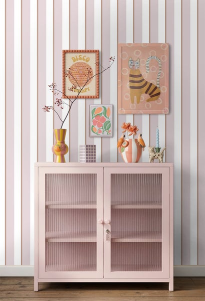 Rosa Vitrinenschrank mit Glastüren vor einer gestreiften Tapete, dekoriert mit bunten Vasen und Bildern in einem hellen Zimmer.