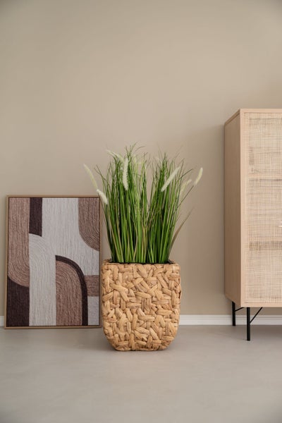 Künstliches Gras mit weißen Wedeln in einem quadratischen geflochtenen Übertopf aus Naturmaterial in einem modern eingerichteten Raum.