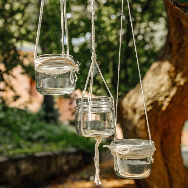 Drei hängende Teelichthalter aus Glas an weißen Seilen in einem hellen Garten.