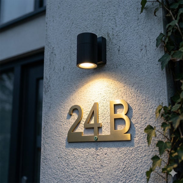 Moderne schwarze Außenwandleuchte beleuchtet eine goldfarbene Hausnummer auf einer strukturierten Fassade.